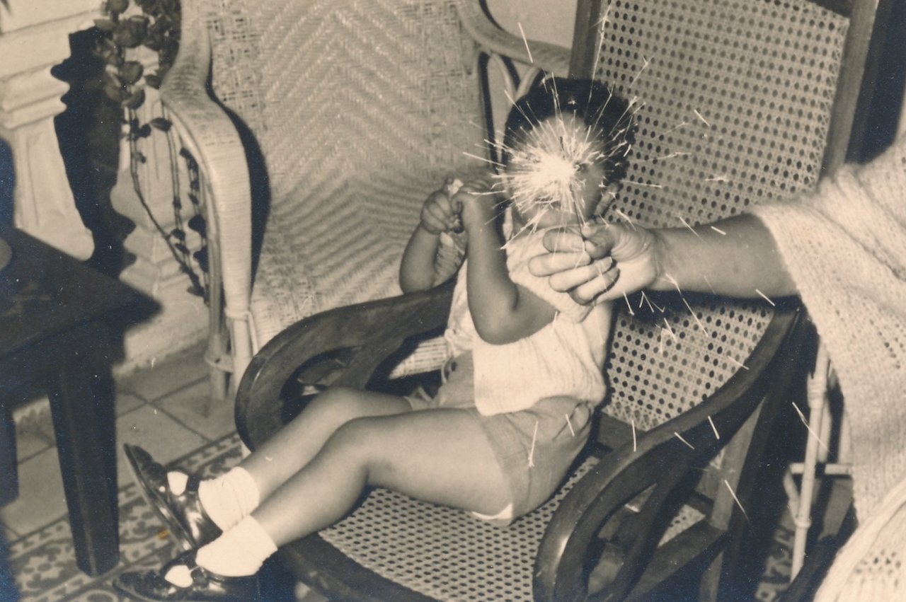 Child holding a sparkler with assistance, sitting on a woven chair indoors.